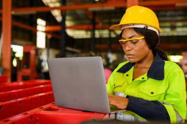 Portrait of female mechanical engineer worker in yellow hard hat and safety uniform using laptop standing at manufacturing area of industrial factory