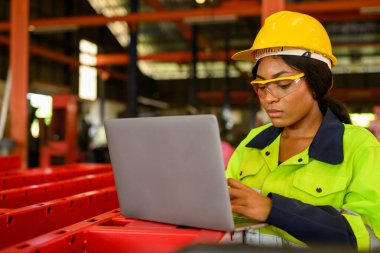 Portrait of female mechanical engineer worker in yellow hard hat and safety uniform using laptop standing at manufacturing area of industrial factory