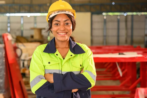 Happy female industrial engineer worker in yellow hard hat standing at manufacturing area posing to camera with arm crossed at industrial factory