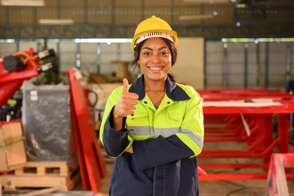 Portrait of happy female industrial engineer worker in yellow hard hat standing at manufacturing area posing to camera with arm crossed at industrial factory