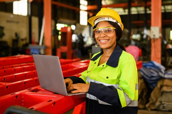 Portrait of female mechanical engineer worker in yellow hard hat and safety uniform using laptop standing at manufacturing area of industrial factory