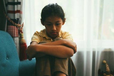 Unhappy girl portrait, Sad child sitting on sofa at home, Upset girl hugging knees alone, Concept of lonely girl and kid with trouble and violence