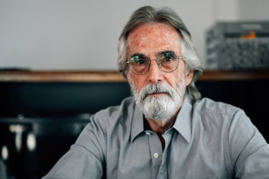 Portrait of senior man with grey hair and beard sitting on sofa at home looking at camera, Healthy senior retirement grandfather resting at home