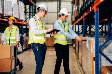 Smart male engineer manager wearing safety vest and helmet using digital tablet checking and assigning work to warehouse workers, Smart teamwork working concept
