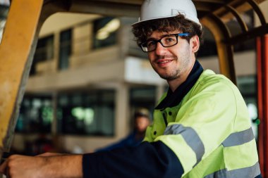 Male industrial engineer in white hard hat driving forklift at manufacturing area working at industrial factory