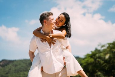 Young happy couple man and woman in white clothes on beach portrait, Girlfriend getting piggyback ride from boyfriend at sunrise over sea beach ocean outdoor in summer day