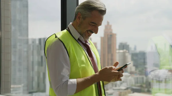 Portrait of happy civil engineer foreman with safety vest using smartphone during break time on construction site, Male contractor enjoying working on building talking with worker team on mobile phone