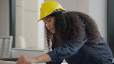 Hardworking female architect with yellow helmet drawing on blueprint of architectural project plan on building site using computer laptop working on high building, Construction work concept