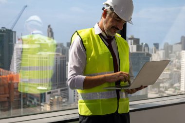 Close up view of male engineer with safety vest holding computer laptop working on construction site, Professional technician typing on laptop working at new building of real estate project
