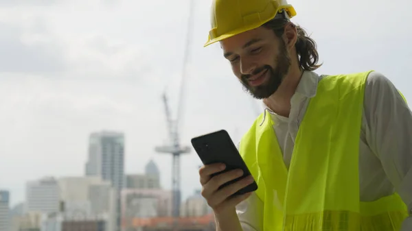 Portrait of smart engineer foreman with safety helmet and vest using smartphone during break time on construction site, Young male architect worker enjoying working on new building