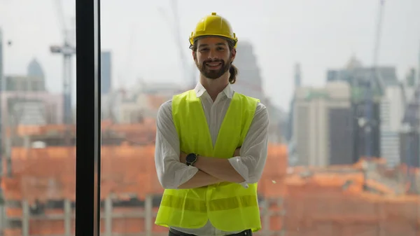 Smart male architect with arms crossed smiling posing to camera, Portrait of confident engineer foreman with safety helmet and vest on building site, Successful contractor smiling happily at workplace