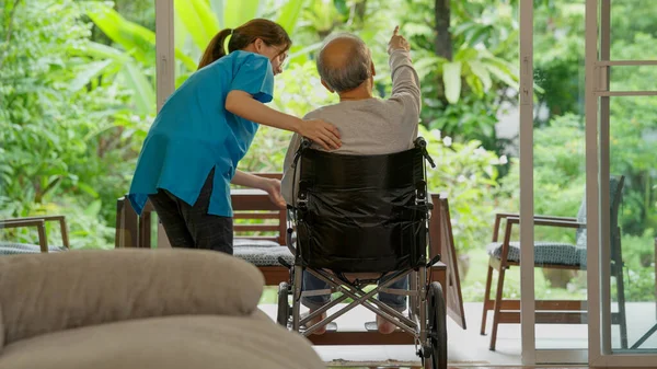 Senior man with knee problems, Elderly patient on wheelchair trying to stand and walk exercising with caregiver nurse at home, Young physiotherapist taking care and supporting senior man to exercise