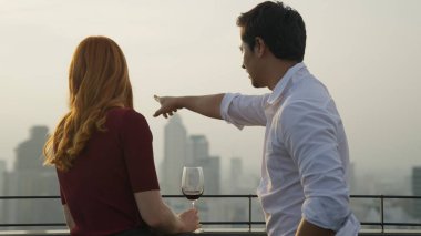 Şarabın neşe kaynağı yüksek bina manzarası günbatımı sıcak tonu ile gökyüzü, mutlu çift cam tutuyor, şarap neşe içinde şehir manzarası, şarap şehrin gökyüzü çatısı manzarası