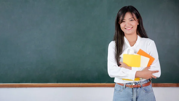 Happy teacher in the classroom, Teacher standing near backboard holding textbook posing to camera with smile, Teacher portrait, Education concept