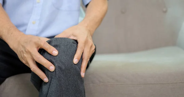 Close up shot of senior man suffering from knee pain sitting on sofa at home, Elderly man touching knee at pain point, People with health problem concept