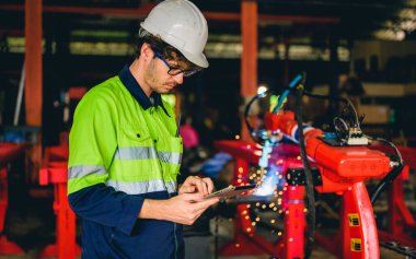 Portrait of happy male mechanical engineer worker in white hard hat and safety uniform standing at manufacturing area holding laptop at industrial factory