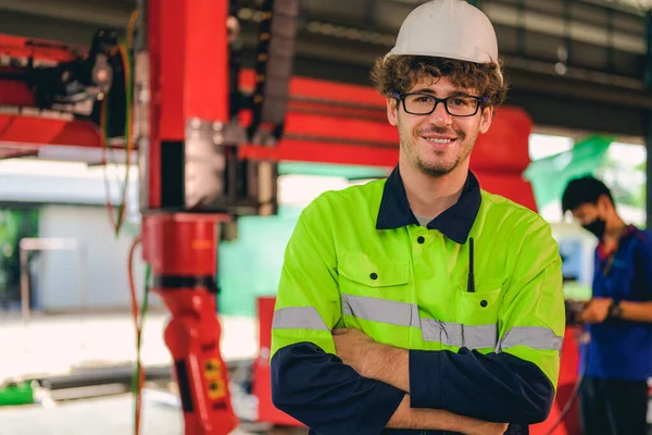 Portrait of happy male mechanical engineer worker in white hard hat and safety uniform standing at manufacturing area holding laptop at industrial factory