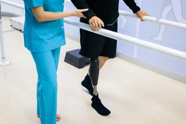 Physiotherapist helping patient with prosthetic leg walk between parallel bars during physiotherapy session at hospital, New artificial limb production for disabled people