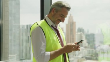 İnşaat sahasında akıllı telefon kullanan güvenlik yeleği olan mutlu inşaat mühendisi portresi, inşaat işçisi cep telefonuyla çalışan işçi ekibiyle konuşmaktan keyif alıyor.