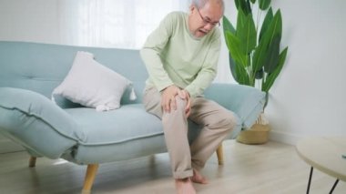 Senior man using hand touching knee at pain point, Elderly man suffering from knee pain sitting on sofa at home, People with health problem concept