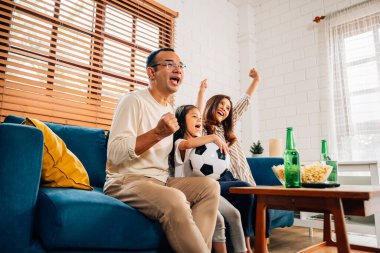 Mutlu bir aile televizyonda maç seyrediyor, insanlar tezahürat yapıyor ve el kaldırıyor, anne ve çocuk evde eğleniyor.