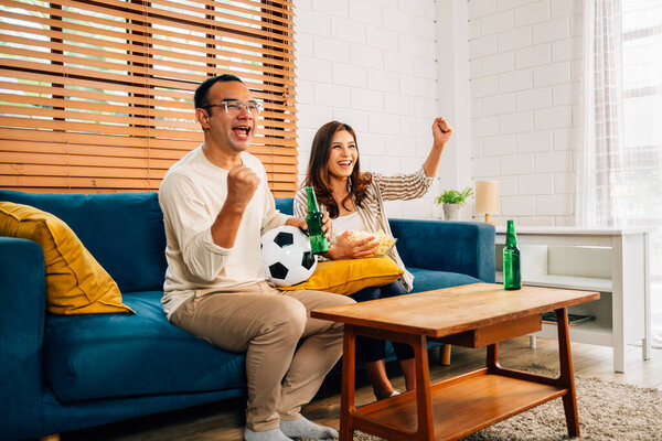 Couple of happy husband and wife watching sports match together on television, People cheering and shouting goal with hands up at living room, Family having fun with activities at home