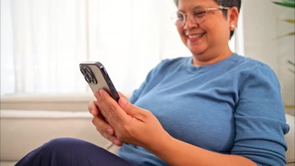 モバイルアプリを使ってスマホを持って友達とおしゃべりする健康な熟女 自宅でテクノロジーを持つシニア 自宅で幸せをもってリラックスするおばあちゃん 高品質の写真 — ストック動画