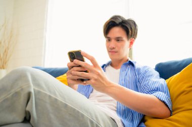 Genç bir adamın cep telefonu kullanmasının ve oturma odasındaki koltukta oturmasının portresi hafta sonları evdeki koltukta oturup akıllı telefon kullanmasının, insanların yaşam tarzı. Yüksek kalite fotoğraf
