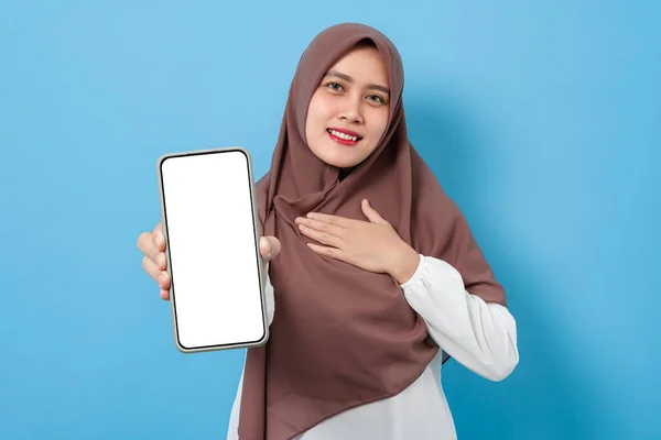 Happy smiling muslim woman showing blank smartphone screen for mock up isolated on blue background. mobile phone mockup application recommendation concept