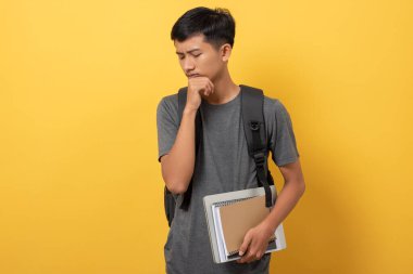 Sırt çantası ve kitaplarıyla gülümseyen genç üniversite öğrencisi sarı arka planda izole edilmiş baş ağrısı ve stresinden muzdarip.