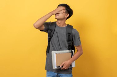 Sırt çantası ve kitaplarıyla gülümseyen genç üniversite öğrencisi sarı arka planda izole edilmiş baş ağrısı ve stresinden muzdarip.