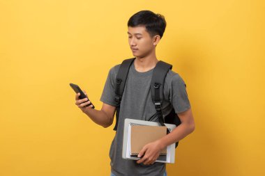 Sırt çantası ve kitaplarıyla gülümseyen genç üniversite öğrencisi sarı arka planda cep telefonu kontrolü yaparak sosyal sağlığını kontrol ediyor.