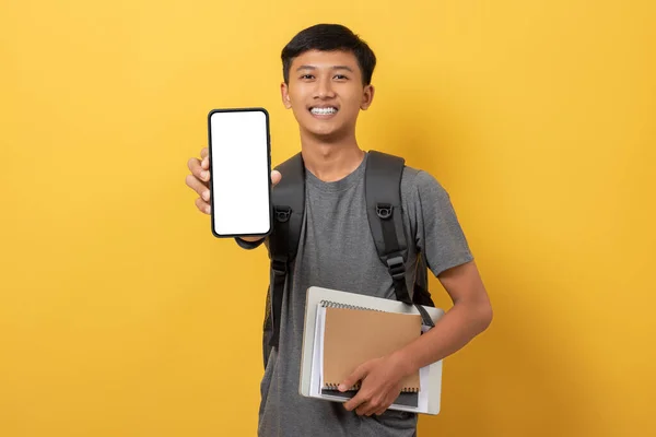 Smiling young college student with books and backpack isolated on yellow background showing big smartphone with white blank screen in hand. Advertisement special offer Concept.