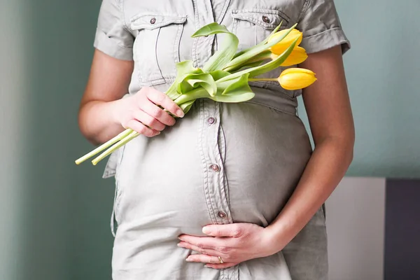 Close up of a pregnant young woman with tulips flowers holding hands on belly on a pastel background. Mothers day, womens day, motherhood or pregnancy concept. High quality photo.