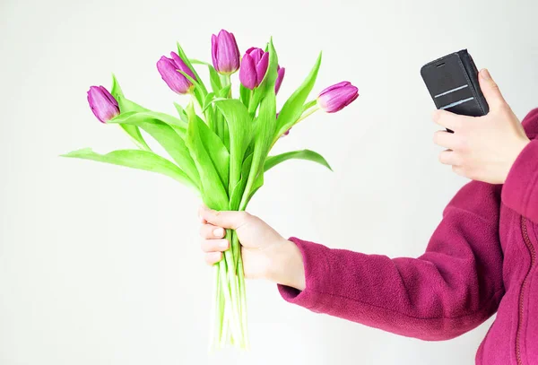 Beautiful girl in cozy purple jacket with magenta tulips is taking a picture from the flowers on her phone. Indoor photo. No face. High quality photo.