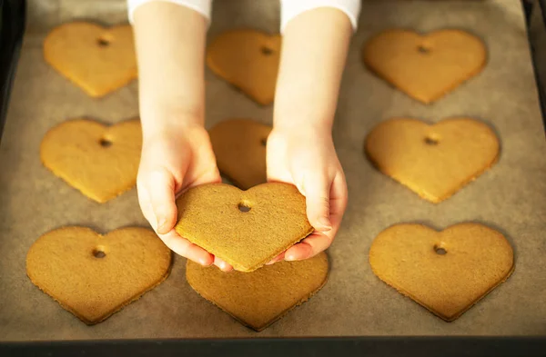 Girl hands holding a heart cookie in front of the homemade fresh baked cookies on baking sheet. Valentines Day concept. High quality photo.