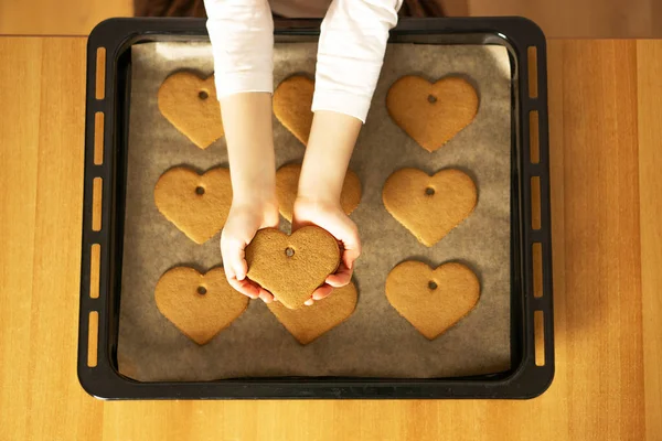 Child hands holding a heart cookie in front of the homemade fresh cookies on baking tray. Valentines Day concept. High quality photo.