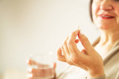 Evinde elinde hap ve cam su tutan günlük kıyafetlerle emekli bir kadının fotoğrafını çek. Sağlıklı, orta yaşlı, elinde bir bardak su ve ilacını tutan bir kadın. Mutlu yetişkin kadın vitamin hapı alıyor..