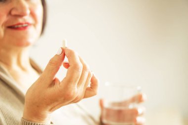 Evinde elinde hap ve cam su tutan günlük kıyafetlerle emekli bir kadının fotoğrafını çek. Sağlıklı, orta yaşlı, elinde bir bardak su ve ilacını tutan bir kadın. Mutlu yetişkin kadın vitamin hapı alıyor..