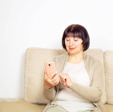 Mutlu, rahat, yaşlı bir kadın evde rahat bir koltukta oturuyor ve akıllı telefon kullanıyor. Gülümseyen yaşlı bayanın portresi cep telefonunu kullanıyor ve internete bağlanıyor. Mesajlaşma, ağ kurma, online