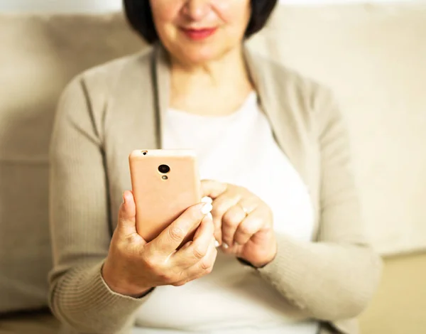 stock image Happy relaxed middle aged woman sitting on comfortable sofa at home and using a smartphone. Portrait of smiling senior lady using her mobile phone and connecting online. Texting, networking, online