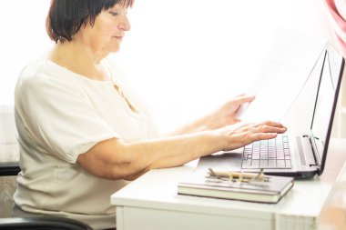 Orta yaşlı yaşlı bir kadın dizüstü bilgisayarı ve kağıt dökümanlarıyla oturmuş, klavyede çalışıyor ve bahşiş veriyor. Elinde kağıt para tutan ve bilgisayar başında çalışan mutlu bir iş kadını. Ev hanımı kıdemli kadın.