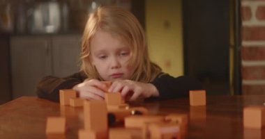 Blond girl plays busily on a table with orange wooden toy blocks, role game with cubes, cute child builds a house for bears. Kids leisure activity. Active imagination. Nice home interior. HQ 4K video