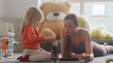 Young sporty mom teaches her daughter with a book on the floor. Cute blond kid, pretty women wearing bright clothes, nursury room filled with natural light. Finding time for sports being a parent. 4K