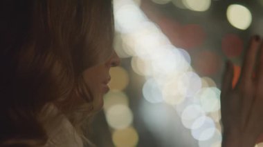 View of tender beautiful girl touches window glass, watches lights of night city in her apartment. Unfocused car lights background, bokeh. Dreaming of somebody loved, waiting and worrying. 4k footage