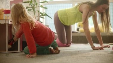 Mother does sport exercises while daughter plays at nursury room, pretty blond kid, women wearing bright clothes, wooden toy house for dolls. Trying to find time for sports being a parent. 4k footage