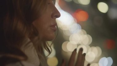 View of tender beautiful woman watches night city lights at window in her bedroom, apartment. Car lights reflected in the glass, bokeh, moving focus. Waiting and worrying about something. 4k footage