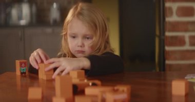Bored sad girl plays busily on a table with orange wooden toy blocks, autistic child, cute kid builds street with houses at kitchen. Mental health problems. Nice home interior. High quality 4K footage