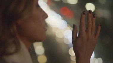 View of tender beautiful woman watches night city lights at window in her apartment, touches glass.Car lights reflected in the glass,bokeh,moving focus.Waiting and dreaming about something.4k footage