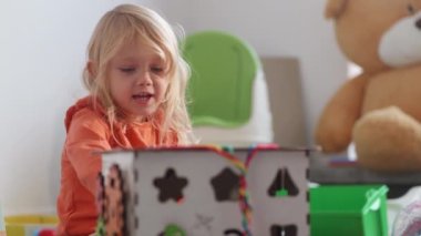 Child plays with busy cube on the floor of nursury room, huge bear on background, than girl takes national culture doll. Educational toys, playtime delight, exploring nationality. HQ 4k footage